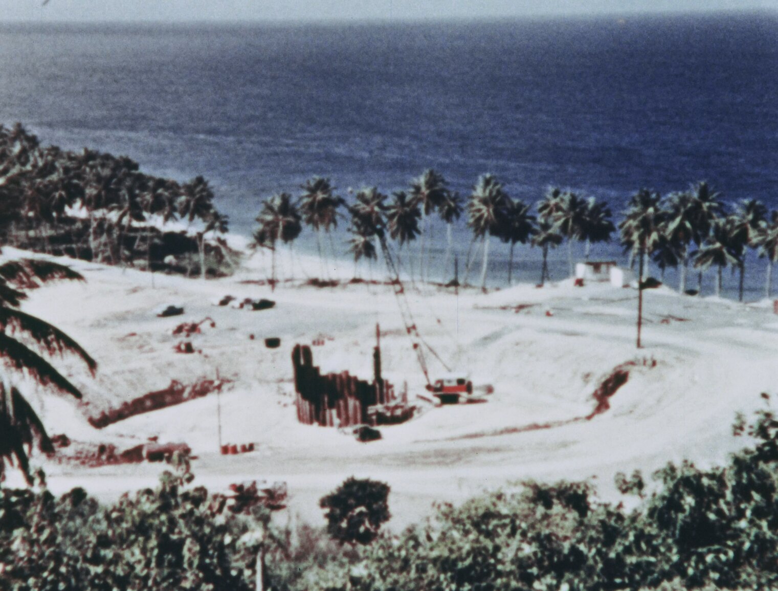 Elevated view of the initial BONUS reactor construction, showing a crane placing steel beams in a cylinder in an excavated area. Palm trees and ocean are seen in the background.