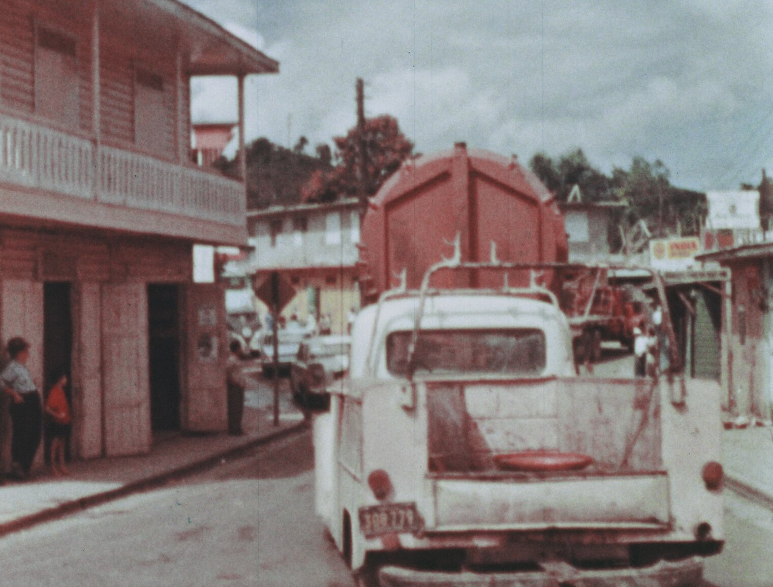 Convoy of trucks carrying the BONUS reactor vessel through a town in Puerto Rico. Adults in hats and barefoot kids stand by watching. Several two-story buildings are seen.