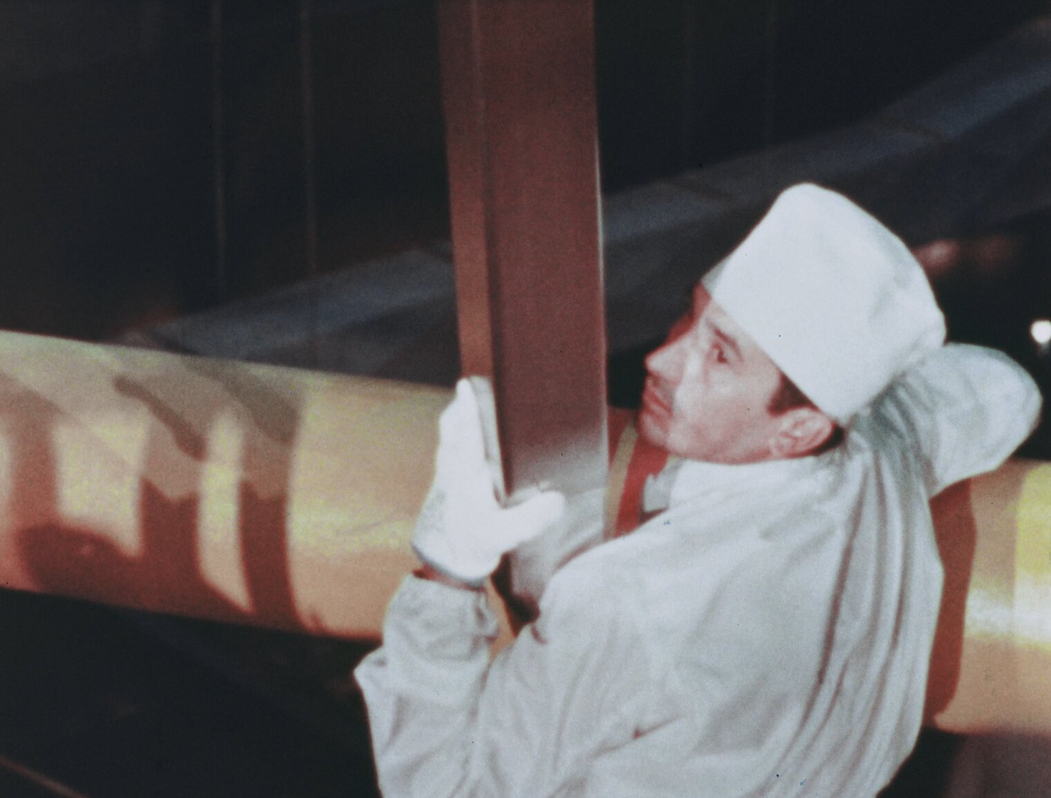 Man in white coveralls, a white cap, and white gloves holds a suspended fresh nuclear fuel assembly close while positioning it for initial core loading at BONUS.