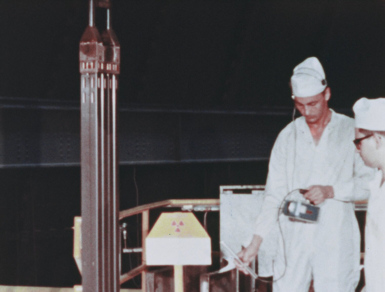 Men in white coveralls and caps look at geiger counter while standing near a nuclear superheat fuel assembly being loaded into BONUS. Dark background. Radiation sign on pedastal.