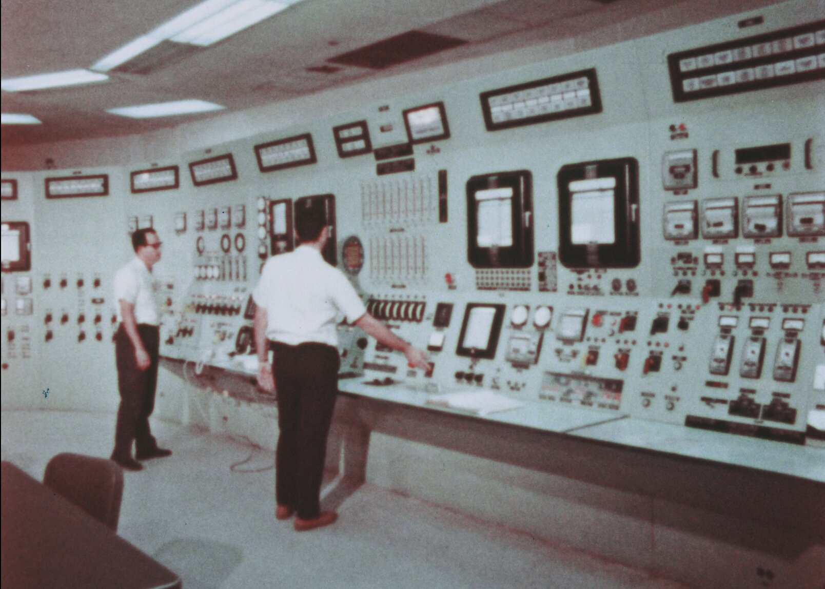 2 Men standing in bonus control room pressing buttons. The overview of the control room is visible in pale green with many indicators, switchces, and buttons, and alarms seen.